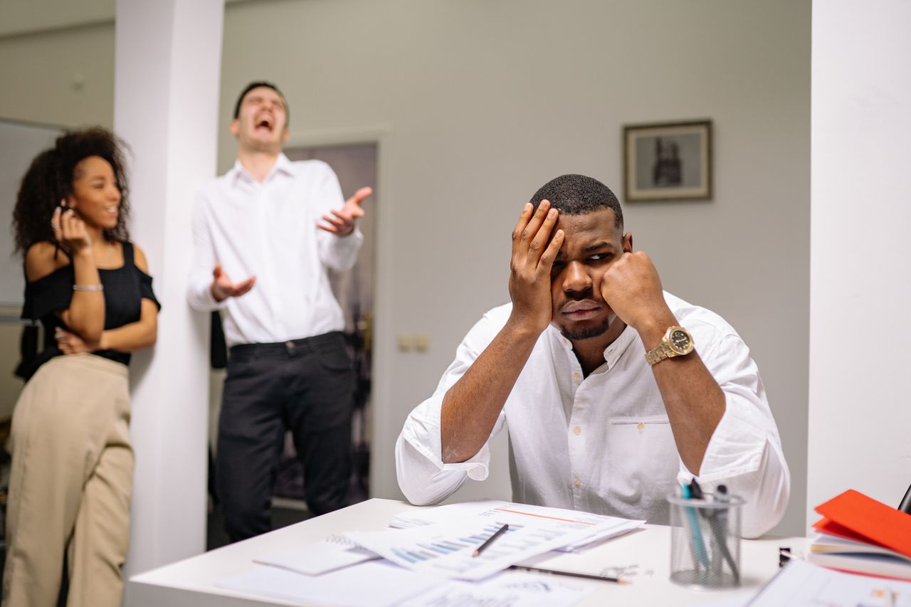 A man at a desk looks frustrated while two coworkers laugh. Office workplace conflict concept.