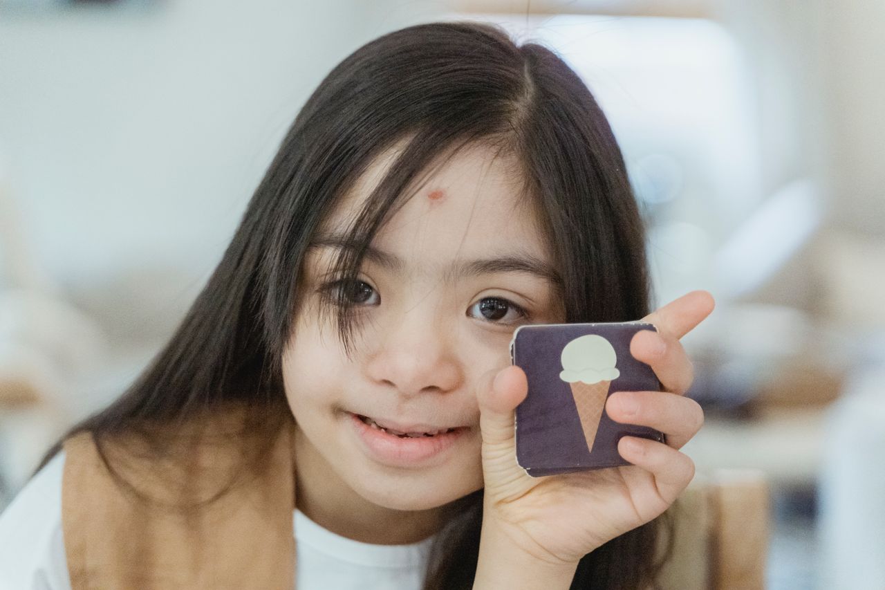 Young girl with Down syndrome smiling and holding a memory game card indoors.